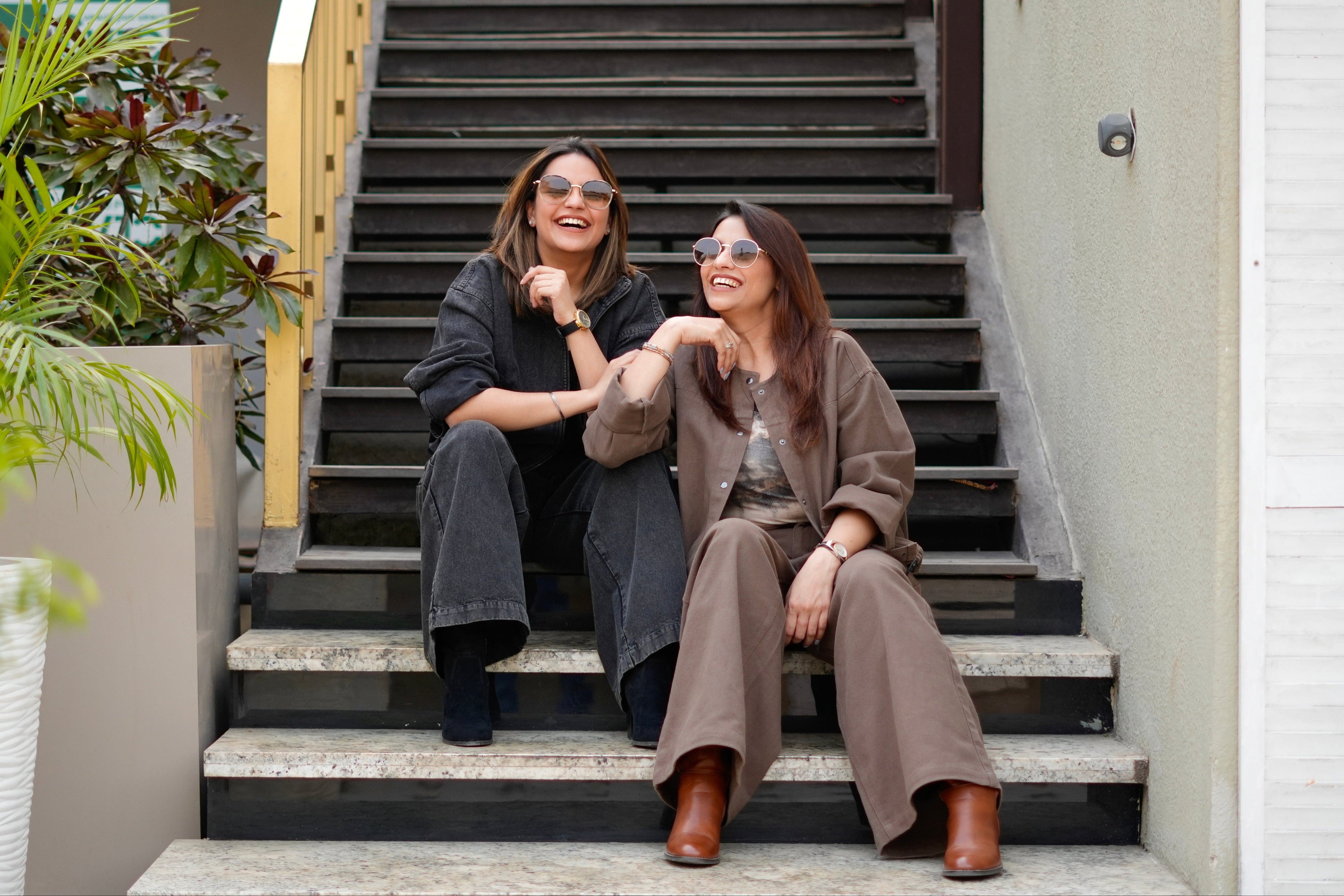 Two women sitting on a set of stairs outdoors, wearing sunglasses and casual clothing.