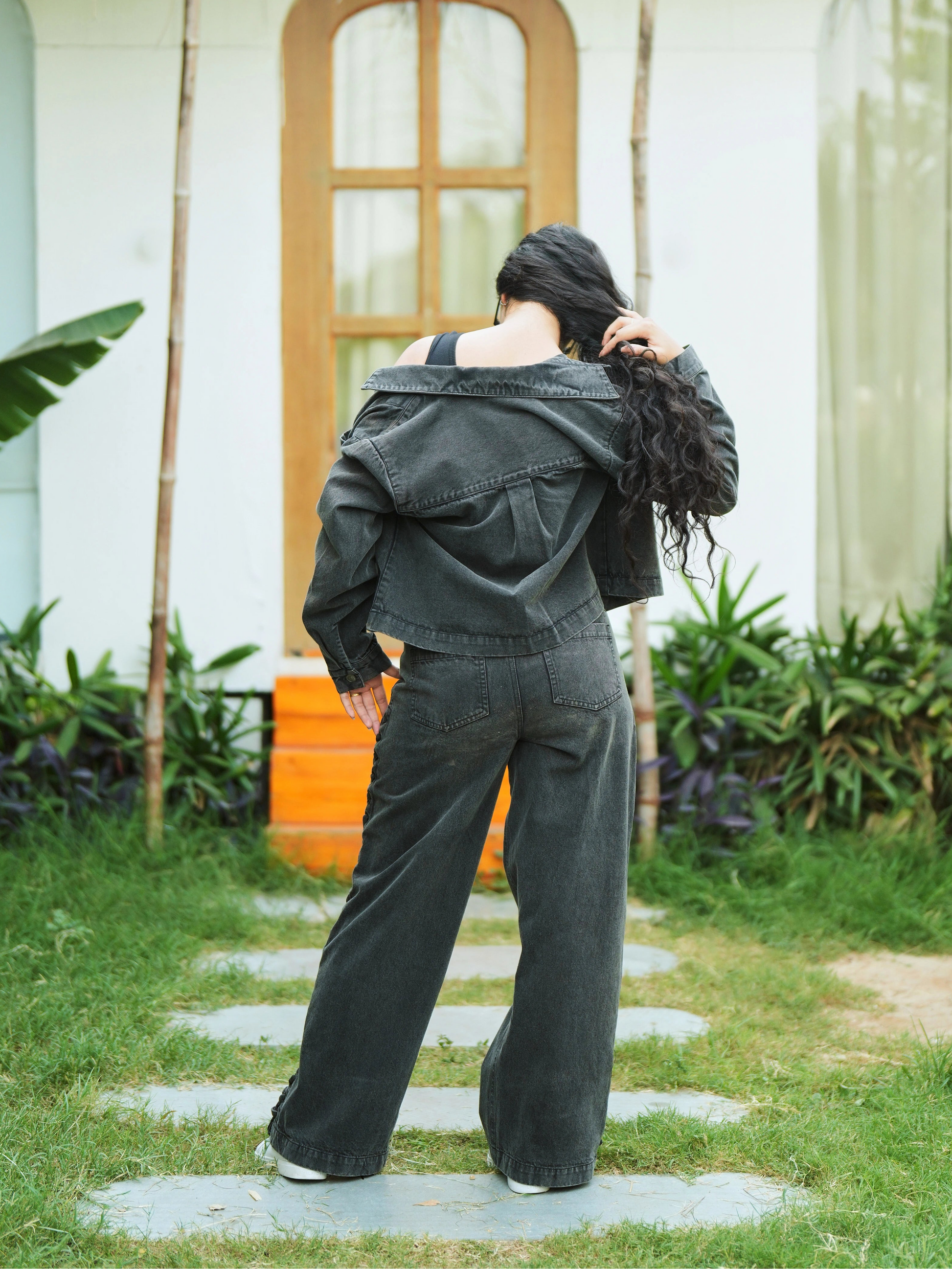 Person in Audela black denim jacket standing on a stone path in a garden with a white building and plants in the background