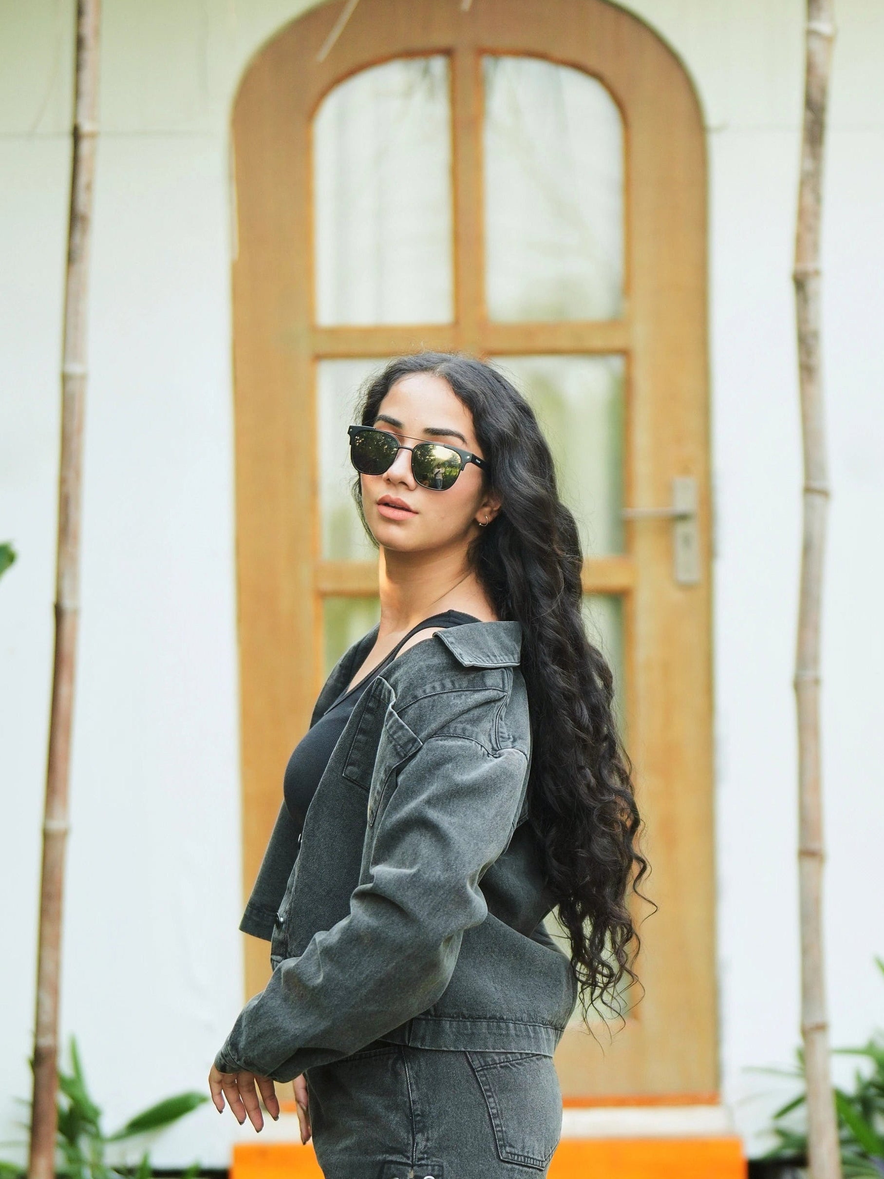 Woman wearing sunglasses and a denim jacket in front of a wooden door.