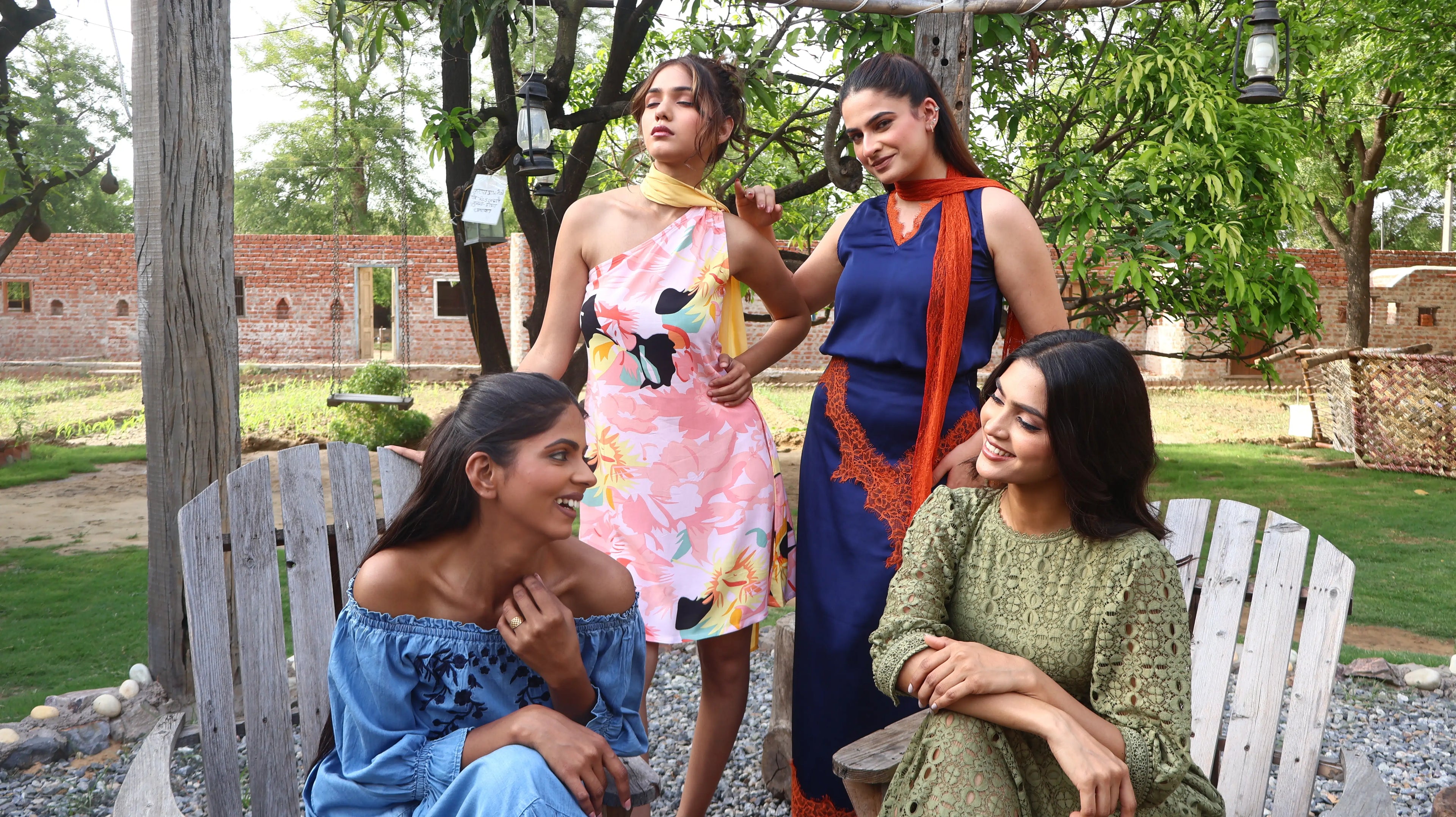 Four women in colorful dresses sitting outdoors on a wooden bench with trees and a building in the background. Audela desktop hero image. Girls' farm stay vacation. 