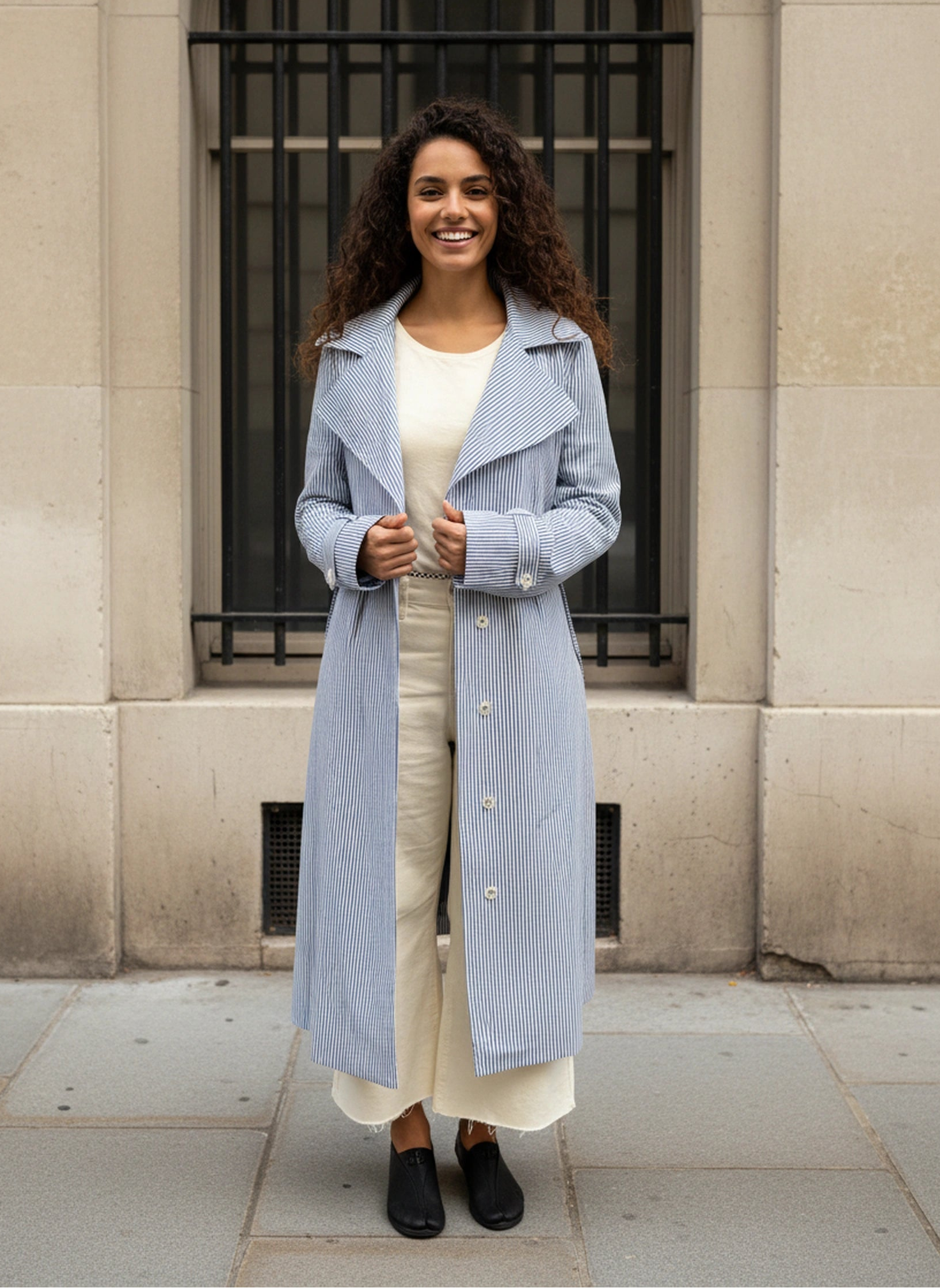 Girl wearing AuDelà long overcoat green, blue, black and white striped. Details on cuff and statement lapels with A-line silhouette. Front button closure. Styled with with t-shirt, white jeans, and black boots