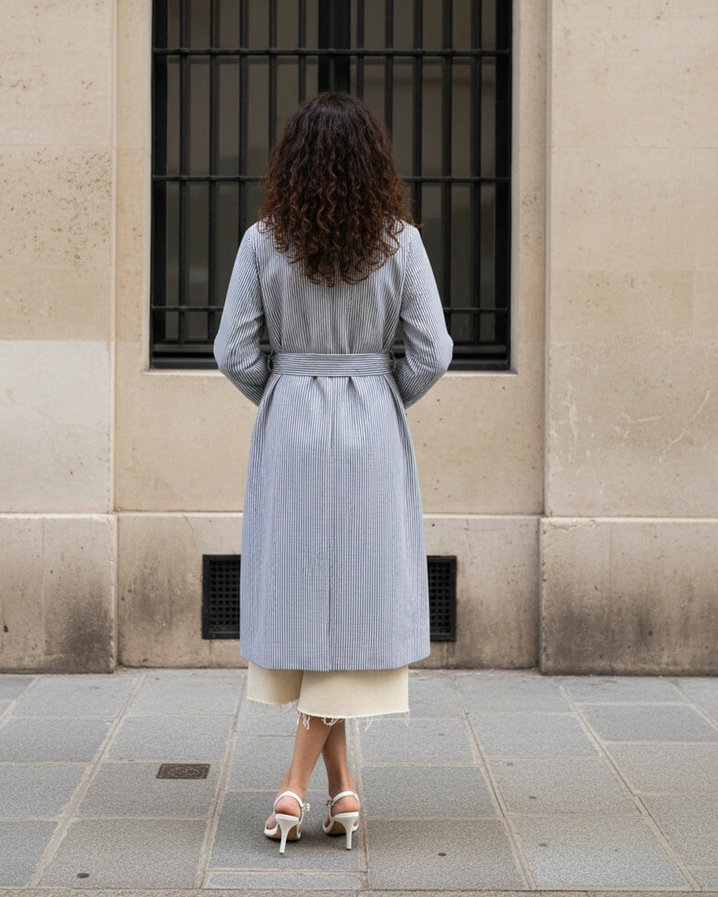 Girl wearing AuDelà long overcoat green, blue, black and white striped. Details on cuff and statement lapels with A-line silhouette. Front button closure. Styled with with t-shirt, white jeans, and black boots. back pose.