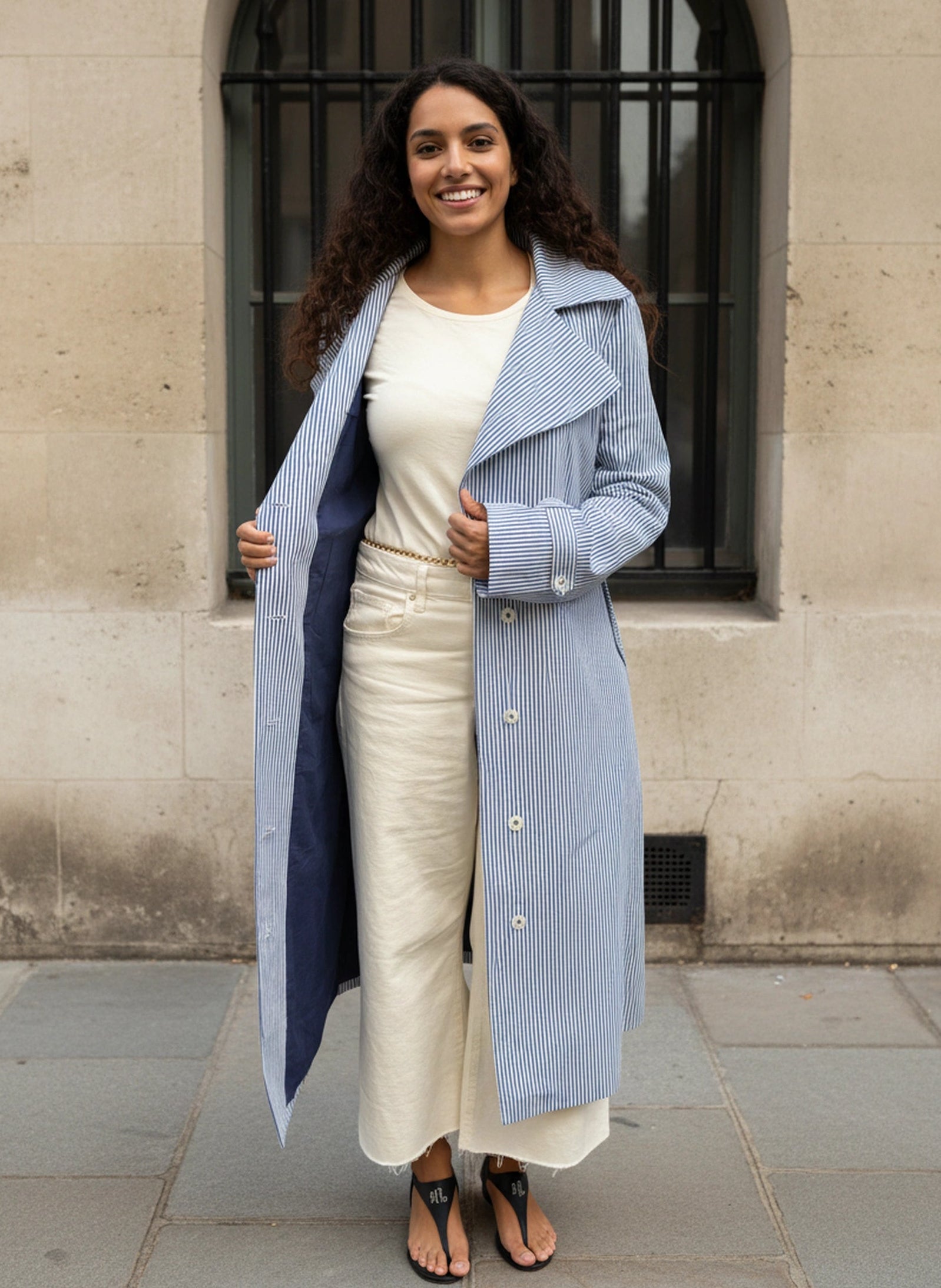 Girl wearing AuDelà long overcoat green, blue, black and white striped. Solid color lining underneath. Details on cuff and statement lapels with A-line silhouette. Front button closure. Styled with with t-shirt, white jeans, and black boots