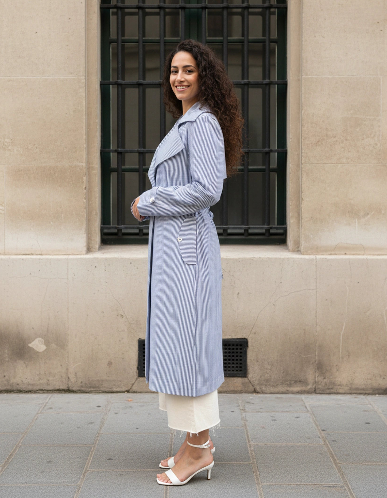 Girl wearing AuDelà long overcoat green, blue, black and white striped. Details on cuff and statement lapels with A-line silhouette. Front button closure. Styled with with t-shirt, white jeans, and black boots. side pose showing side pockets.