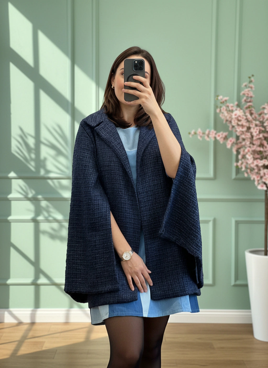 Person wearing a blue coat over a light blue dress, standing in a room with a green wall and a plant taking a full view mirroe selfie