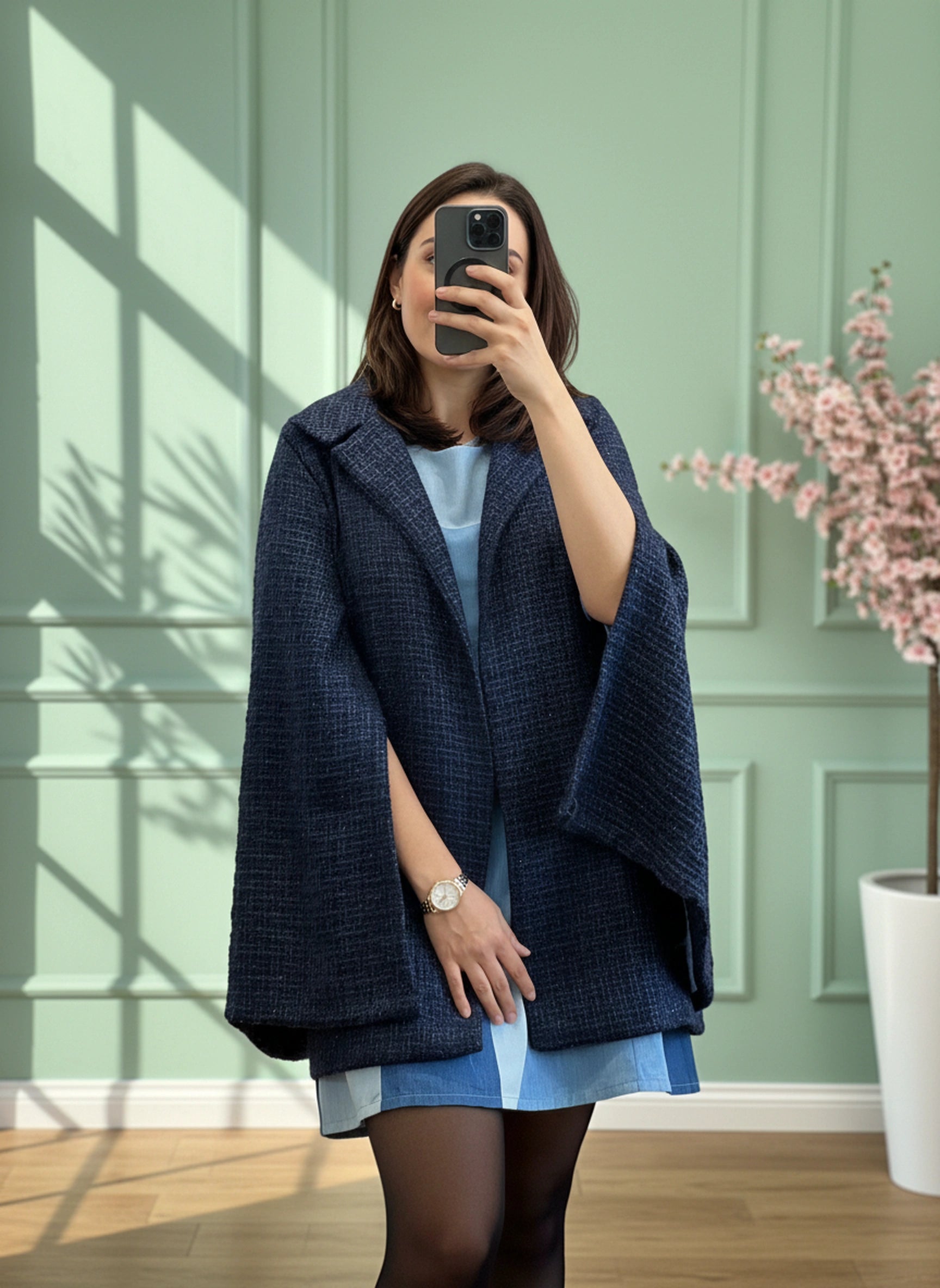 Person wearing a blue coat over a light blue dress, standing in a room with a green wall and a plant taking a full view mirroe selfie